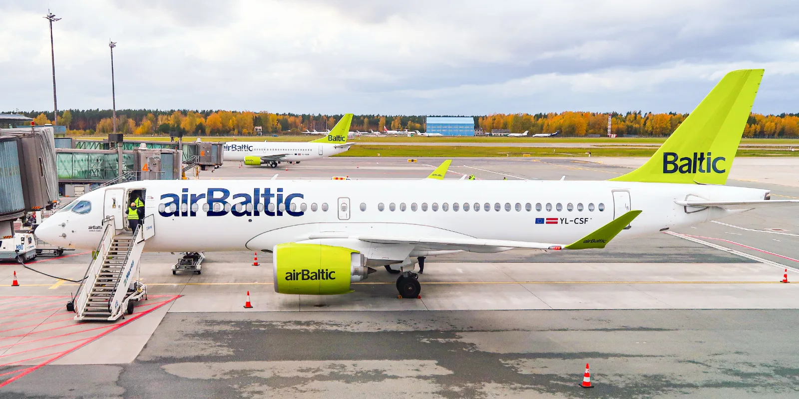 airBalticu valge-roheline Airbus A220-300 seisab Riia lennujaama perroonil, uksel trepp ja taustal sügisesed puud