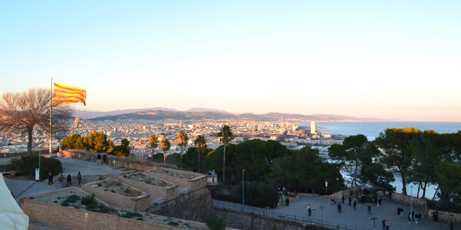 Vaade Montjuïci kindluse vallilt Barcelona kesklinnale ja Vahemerele, esiplaanil Kataloonia lipp, männid ja palmid, taamal mäed
