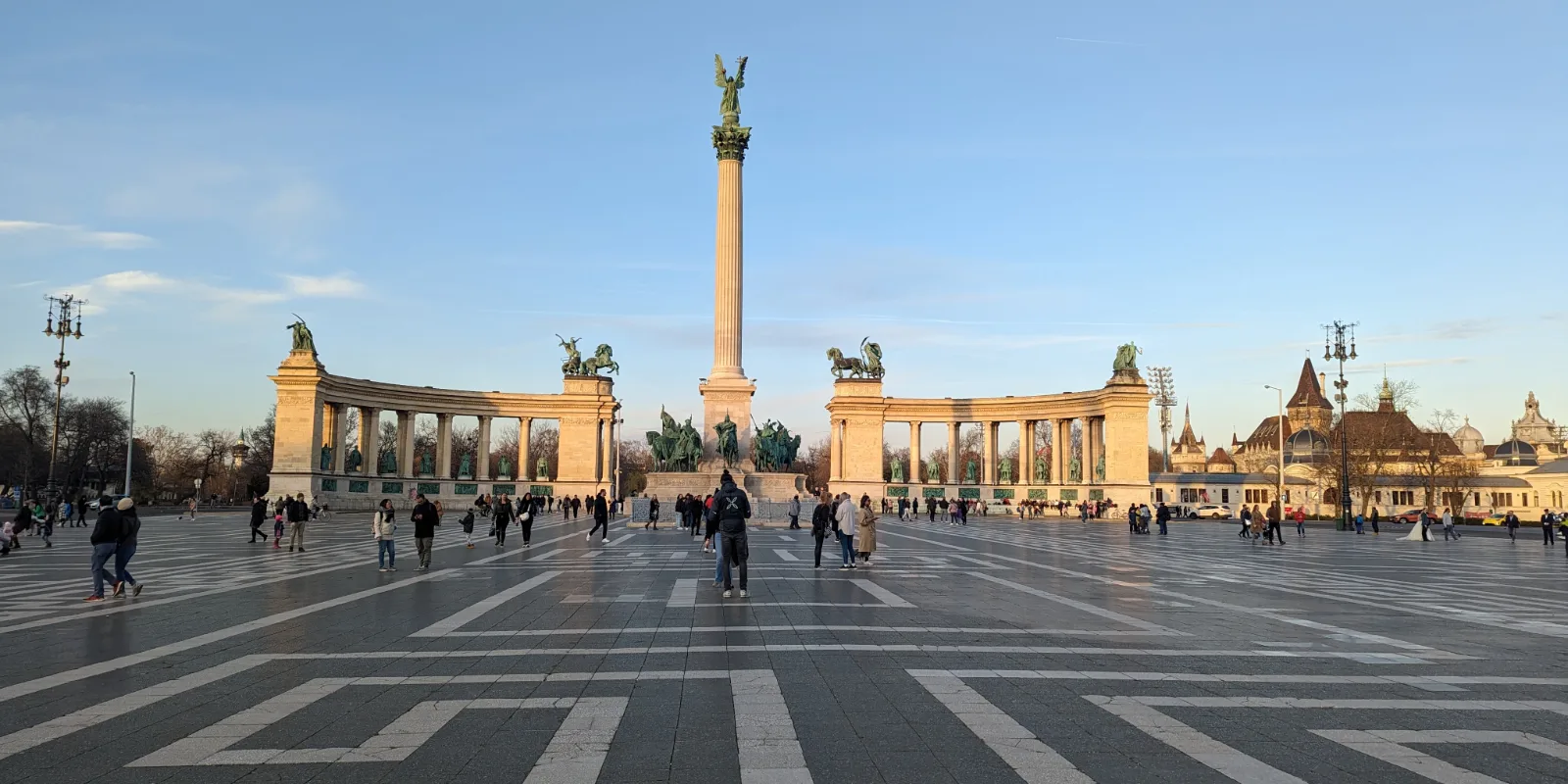 Budapesti Kangelaste väljak madalas päikesevalguses: Milleniumi sammas ingliskujuga, kaarjad sambakäigud ja mustriline kiviparkett, vasakul raagus puud