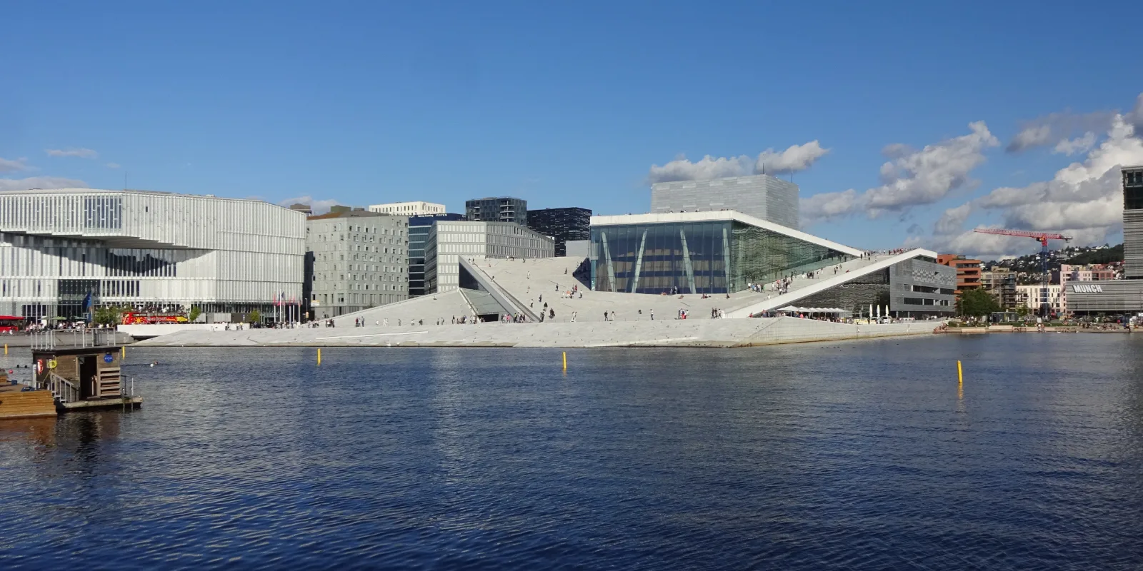 Oslo ooperimaja valge viltune katus vee ääres, taga Barcode-kvartali kõrghooned ja sinine taevas