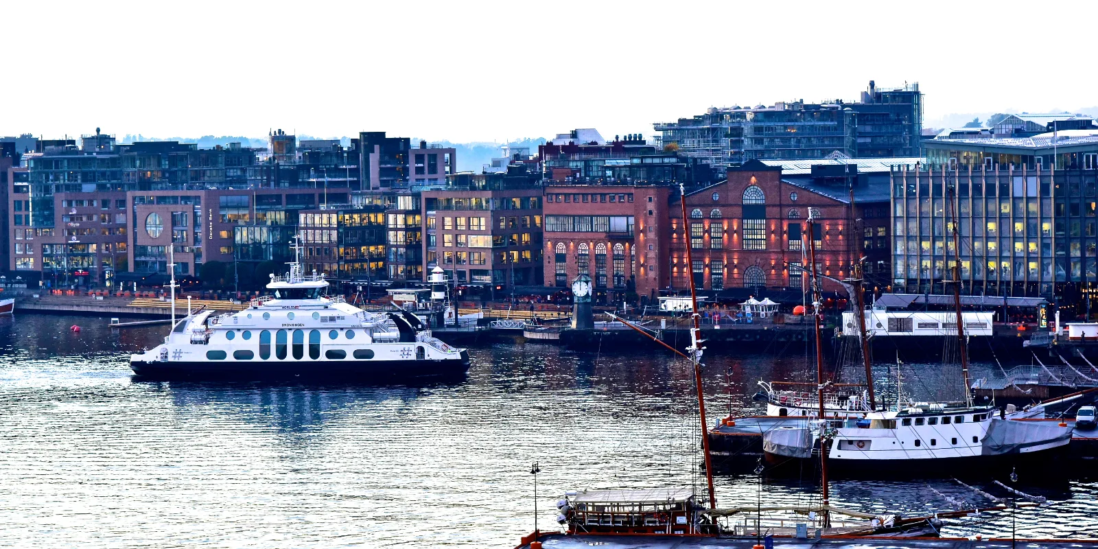 Oslo sadam hämaras: valgustatud akendega telliskivihooned kaldal, valge parvlaev ja purjelaevad vee peal